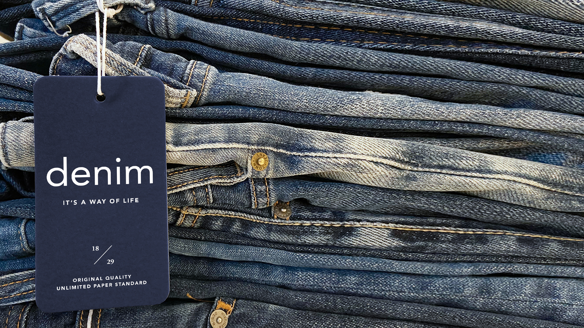 Aufeinander gelegte Jeanshosen. Ein plastikfreies Papierhangtag mit weißem Faden ist an einer Gürtelschnalle befestigt